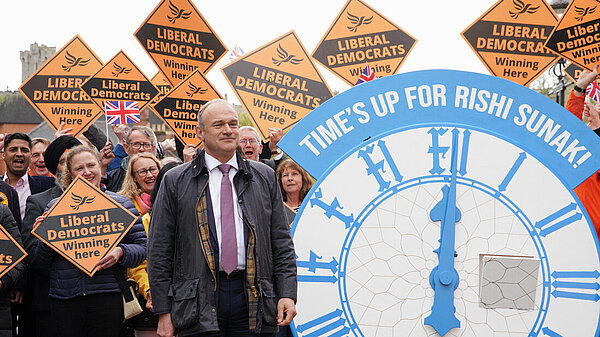 Ed Davey in front of crowd with Lib Dem diamond signs and clock with words time's up for Rishi Sunak